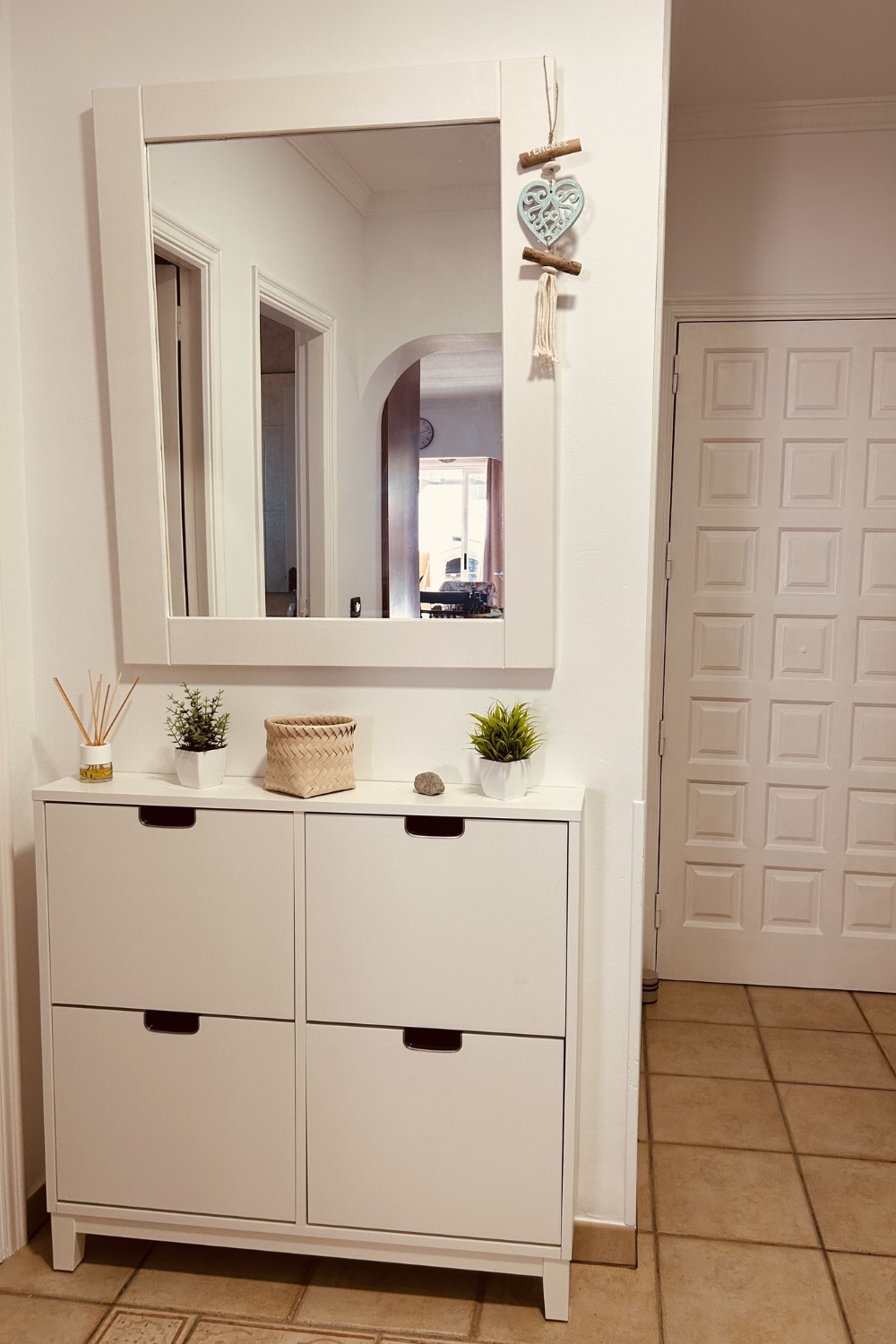 A white mirrored console table with a wall mirror and small plants in a quiet entryway